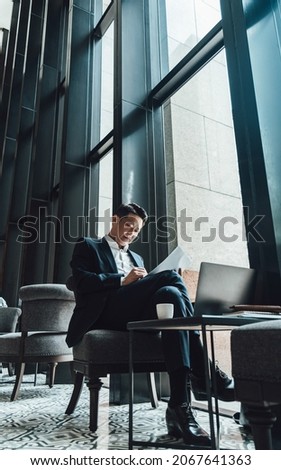 Successful Asian Businessman Working from a Cafe. 
Low angle view of serious business man in blue suit writing something on a paper while sitting in a cafe next to the big windows. Royalty-Free Stock Photo #2067641363