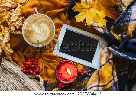 Autumn or winter home cozy composition with yellow scarf, checkered plaid, cup of coffee, dried leaves, burning candle and e-book with copy space. Fall season template for feminine blog social media.