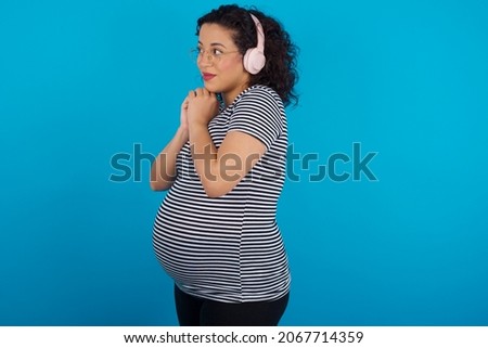 Young pregnant arab woman wearing striped T-shirt on blue studio background wears stereo headphones listening to music concentrated and looking aside with interest.