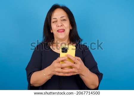 Pleased middle aged Arab woman standing against blue background using self phone and looking and winking at the camera. Flirt and coquettish concept.