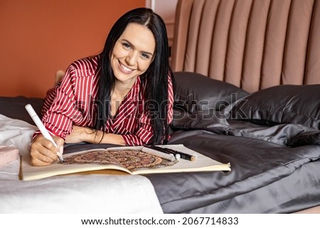 Happy young brunette woman in pajama lying on bed painting antistress picture with colorful pencil having positive emotion. Smiling relaxed pleasant feminine spending time to art therapy hobby