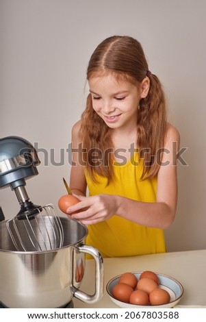 A positive girl of 9-10 years cooks homemade cake in the kitchen, beats eggs in a mixer on the kitchen table. Against the background of a white-gray wall. close-up. High quality photo