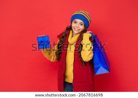 happy kid in winter hat hold shopping bag and gift box on red background, purchase