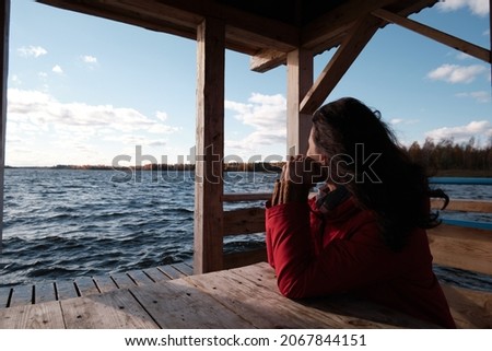 A woman in a Red Jacket Sits at a Table on a Wooden Pier and Looks at the Water, Clouds and Trees with Yellow Foliage. Autumn Landscape.