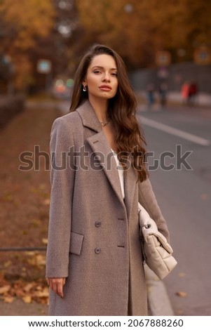 Elegant brunette woman with long wavy hair wearing grey wool coat, holding white handbag and walking at city street on autumn day. Portrait against yellow leaves Royalty-Free Stock Photo #2067888260