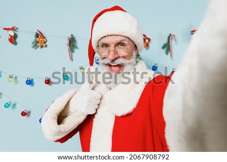 Close up old bearded Santa Claus man 50s in Christmas hat red suit do selfie shot pov on mobile phone show thumb up isolated on plain blue background studio Happy New Year 2022 merry ho x-mas concept