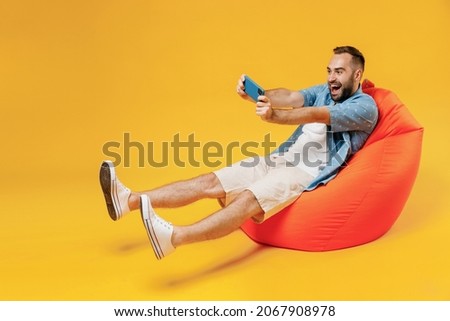 Full body gambling young man in blue shirt white t-shirt sit in bag chair using play racing app on mobile cell phone hold gadget smartphone for pc video game isolated on plain yellow background studio