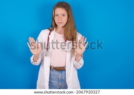 Serious Young caucasian doctor woman wearing medical uniform and stethoscope over blue background pulls palms towards camera, makes stop gesture, asks to control your emotions and not be nervous