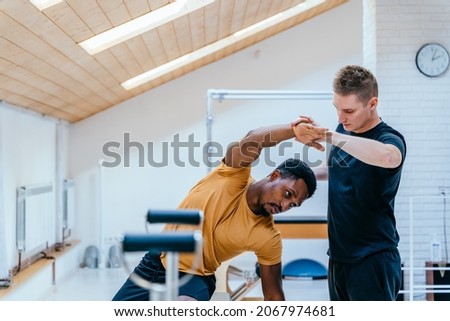 African american man doing exercise on the pilates chair, supporting and helping his positive caucasian man instructor, personal trainer. Special exercise programs for people concept. Royalty-Free Stock Photo #2067974681