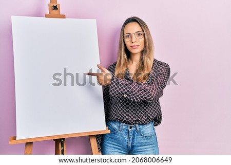 Beautiful hispanic woman pointing to painter easel stand relaxed with serious expression on face. simple and natural looking at the camera. 