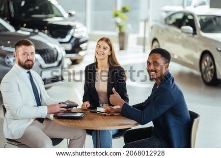 Multi ethnic couple and salesman sitting together at table and making contract about car purchasing. African man and caucasian woman paying with credit card at dealership center.