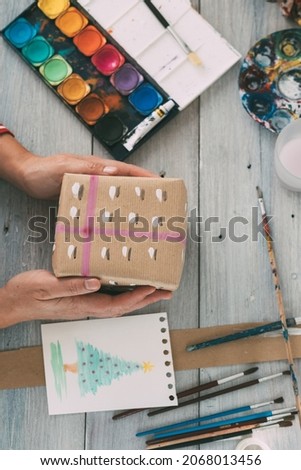 Person holding the gift wrapped with watercolor painted paper next to the drawing of a Christmas tree, the brushes and watercolor on the table