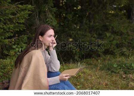 Young woman drawing with pencil in sketchbook near forest, space for text