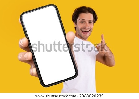 Cheerful young guy holding smartphone with white empty screen, pointing at blank display for mobile application advertisement or your text, gadget mockup template, isolated orange studio background