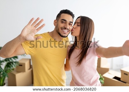 Lovely Caucasian woman kissing her Arab boyfriend, taking selfie, moving to new home together. Loving young multiracial couple photographing themselves on first day in rented apartment