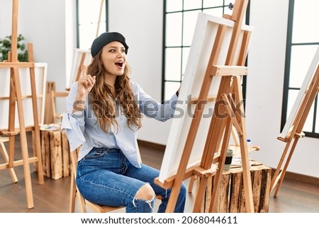 Young hispanic artist woman painting on canvas at art studio pointing finger up with successful idea. exited and happy. number one. 