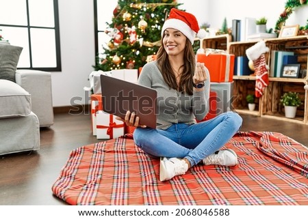Young latin woman using laptop sitting by christmas tree doing money gesture with hands, asking for salary payment, millionaire business 