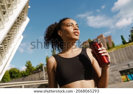 Black sportswoman drinking water while working out on parking outdoors