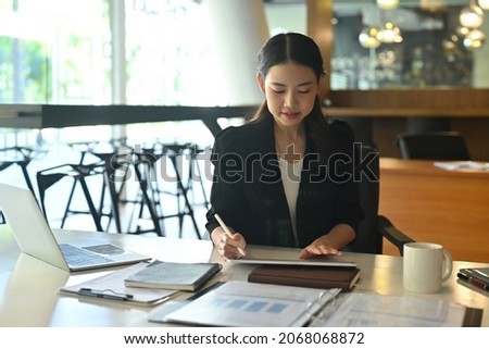 Photo of a businesswoman using a digital tablet and stylus pen at the modern working desk surrounded by a computer laptop, coffee cup and document.