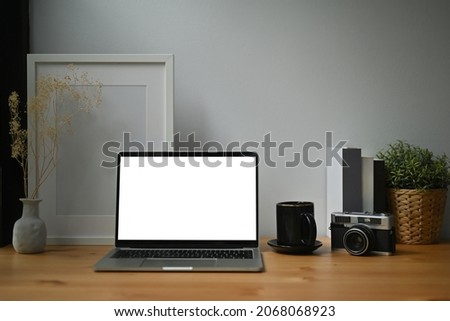 Photo of a white blank screen computer laptop on the wooden table surrounded by a coffee cup, vintage camera, empty picture frame potted plant and books.