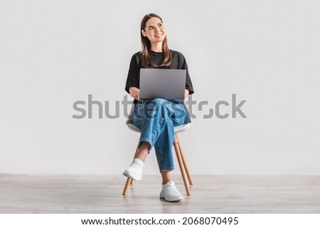 Beautiful young woman sitting on chair, working on laptop against white studio wall, full length. Dreamy millennial lady using portable pc for work, entertainment or online studies Royalty-Free Stock Photo #2068070495