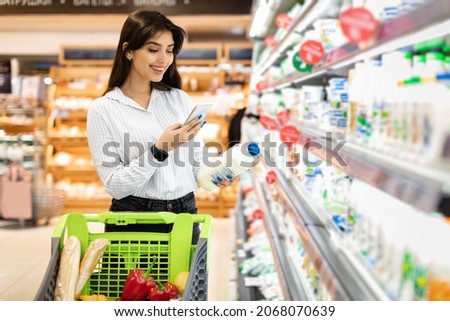 Middle Eastern Woman Using Phone Scanning Milk Bottle Doing Grocery Shopping In Supermarket, Standing With Shop Cart Full Of Products. Consumerism And Technologies, Customer's Application Royalty-Free Stock Photo #2068070639