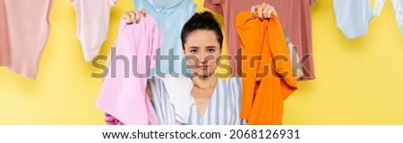housewife looking at camera while holding laundry near clothing hanging on yellow background, banner