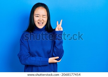 Young chinese girl wearing casual clothes smiling with happy face winking at the camera doing victory sign. number two. 