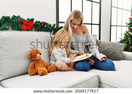 Mother and daughter reading book sitting by christmas tree at home
