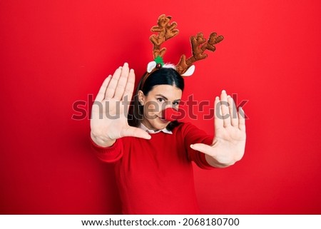 Young hispanic woman wearing deer christmas hat and red nose doing frame using hands palms and fingers, camera perspective 