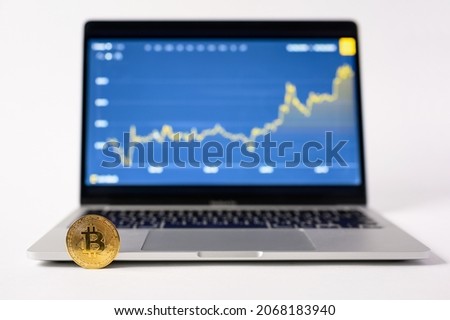 A gold bitcoin coin stands by a laptop against the background of a display with a growing graph