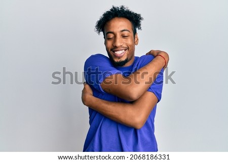 Young african american man with beard wearing casual blue t shirt hugging oneself happy and positive, smiling confident. self love and self care 