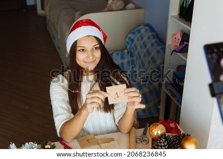 a smiling blogger girl in a Christmas hat makes an advent calendar and takes pictures on her phone