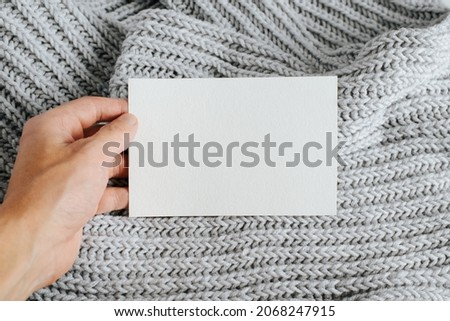 Close-up of hand showing white postcard mockup with blank space for text or bernding. Card template on background of knitted light scarf.