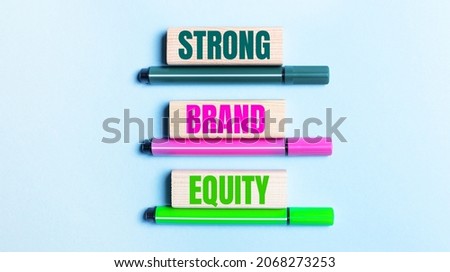 On a light blue background, there are three multi-colored felt-tip pens and wooden blocks with the STRONG BRAND EQUITY