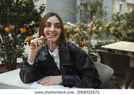 Lifestyle portrait of attractive fashionable brunette pretty woman in eco leather jacket holding pen near lips, smiling looking aside sitting on cafe terrace against the background of tangerine trees