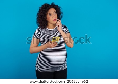 Young pregnant arab woman wearing striped T-shirt on blue studio background thinks deeply about something, uses modern mobile phone, tries to made up good message, keeps index finger near lips.