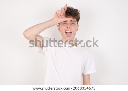 young caucasian man with curly hair wearing T-shirt ​white background gestures with finger on forehead makes loser gesture makes fun of people shows tongue