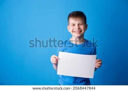 Portrait of a European boy in a blue T-shirt holding a white cardboard on a blue background, a schoolboy smiles and looks at the camera and holds an advertising sheet
