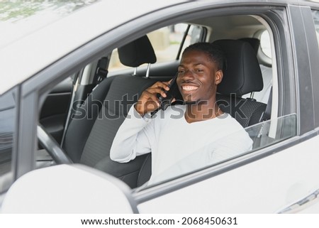 Happy smiling African American male driver sitting behind the self-driving steering wheel of an autonomous electric modern car. Happy guy holds phone and smiles to camera in modern electric car