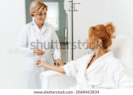 Side view portrait of attractive woman in white bathrobe sitting in armchair with pleasure smile while receiving IV infusion  Royalty-Free Stock Photo #2068463834