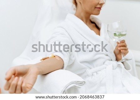 Close up of calm female patient sitting with tube and needle during IV infusion. Girl holding glass with lemon water and relaxing  Royalty-Free Stock Photo #2068463837