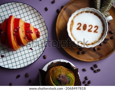 Happy new year 2022 theme coffee cup with number 2022 over frothy surface served on wooden plate with dried pine branches flat lay on purple background with coffee beans, croissant, cruffin (top view)