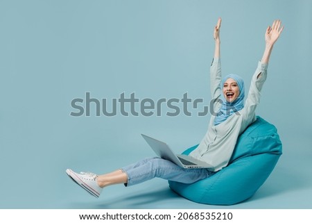 Full body young arabian asian muslim woman in abaya hijab sit in bag chair use work laptop pc computer stretch hands isolated on plain blue background People uae middle eastern islam religious concept Royalty-Free Stock Photo #2068535210