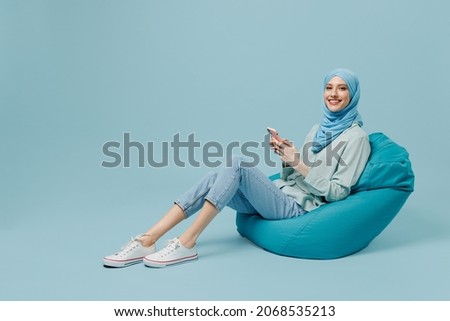 Full body happy young arabian asian muslim woman in abaya hijab sit in bag chair hold use mobile cell phone isolated on plain blue background studio People uae middle eastern islam religious concept Royalty-Free Stock Photo #2068535213