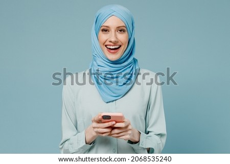 Young amazed happy arabian asian muslim woman in abaya hijab hold in hand use mobile cell phone isolated on plain blue background studio portrait. People uae middle eastern islam religious concept.