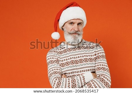 Old aggrieved disconcerted sad upset fatigued tired frowning bearded Santa Claus man 50s wears Christmas hat sweater posing looking camera hold hands crossed isolated on plain orange background studio