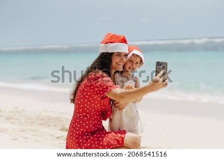 Mother and little aughter in santa hats on the beach making selfie on mobile phone and smiling