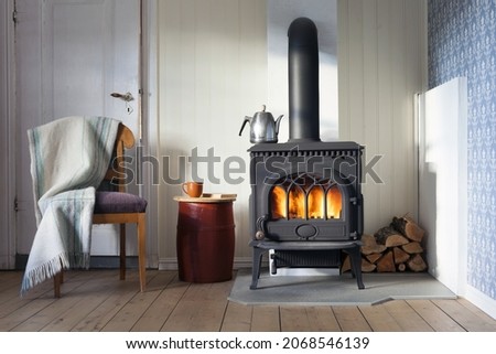 Interior with wood heating stove, chair with plaid and firewood in corner. Red-painted old barrel serving as table for book and coffee cup. Metal coffee pot on stove. Sunlight from outside. Royalty-Free Stock Photo #2068546139
