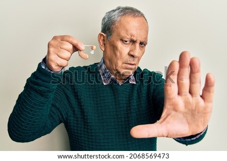 Handsome senior man with grey hair holding medical hearing aid with open hand doing stop sign with serious and confident expression, defense gesture 